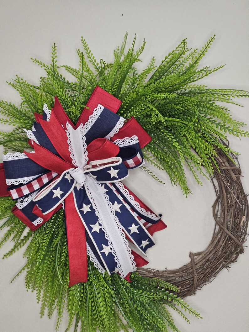 May include: A patriotic wreath featuring a grapevine base, adorned with lush green foliage and a large bow. The bow incorporates red, white, and blue ribbons, some with star patterns and lace trim, creating a festive and decorative piece.