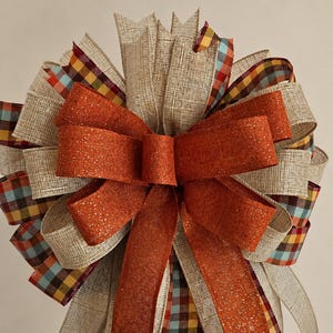 May include: A decorative bow constructed from multiple ribbons. The design features solid orange glittered ribbon, natural tan textured burlap-style ribbon, and a plaid ribbon pattern in shades of orange, yellow, blue, and dark brown.