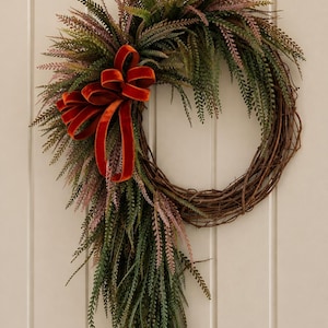 May include: A circular wreath constructed from brown intertwined twigs, adorned with cascading green and muted purple fern-like foliage. A prominent, deep orange velvet ribbon is tied in a multi-looped bow on the left side of the wreath.