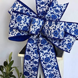 May include: A decorative bow made of white fabric with a blue floral pattern. The ribbon features a repeating design of blue flowers and vines on a white background, with solid blue edges. The bow has multiple loops and two long hanging tails.