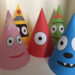 Op de afbeelding: Vijf kleurrijke feesthoeden met cartoongezichten. De hoeden zijn kegelvormig en hebben verschillende ontwerpen, waaronder een roze hoed met een madeliefje, een gele hoed met een robotontwerp, een rode hoed, een groene hoed en een blauwe hoed met een lachend gezicht.