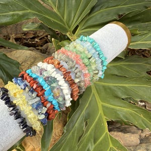 May include: A collection of gemstone chip bracelets in various colors, including blue, yellow, red, and green, are displayed on a white cylindrical stand. The bracelets are made of small, irregular-shaped stones. The background features large green leaves.