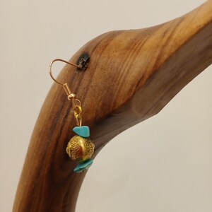 Könnte beinhalten: Ein einzelner goldfarbener Ohrring mit einer strukturierten Perle und türkisfarbenen Akzenten. Der Ohrring hängt an einem goldfarbenen Haken. Der Ohrring wird auf einem Holzständer präsentiert.