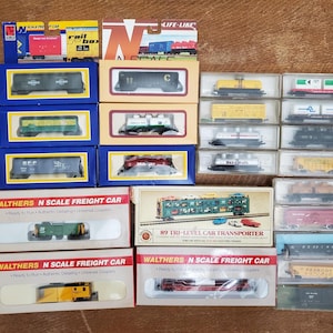 May include: A collection of N scale model train freight cars in various packaging styles, including blue and yellow boxes, white Walthers boxes, and clear plastic cases. The items feature diverse train car types, including tankers, flatbeds, and transporters, with visible branding and model details.