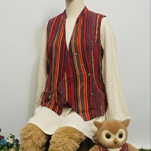 May include: A mannequin wearing a red, yellow, and blue striped vest over a white long-sleeved shirt. The mannequin is also wearing brown furry leg coverings and sitting next to a stuffed owl wearing a matching vest. The owl is sitting on a wooden box.