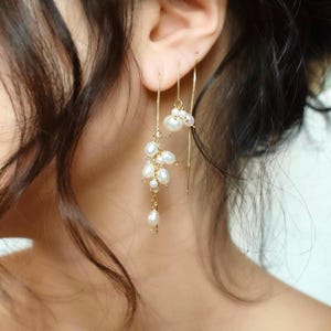 May include: A pair of gold-tone threader earrings featuring clusters of small, white, irregular-shaped freshwater pearls. The earrings consist of thin gold chains that pass through the earlobe, with one side displaying a larger pearl cluster.