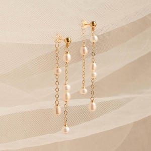 Puede incluir: Un par de pendientes colgantes en tono dorado con múltiples cadenas delicadas y pequeñas perlas de agua dulce de color blanco roto. Cada pendiente consta de dos hilos colgantes de distintas longitudes unidos a un pequeño poste.