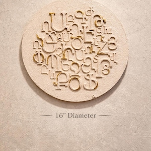 May include: A round, off-white wall art piece with a textured surface. The design features raised, stylized letters with gold glitter accents. The diameter of the art piece is 16 inches.