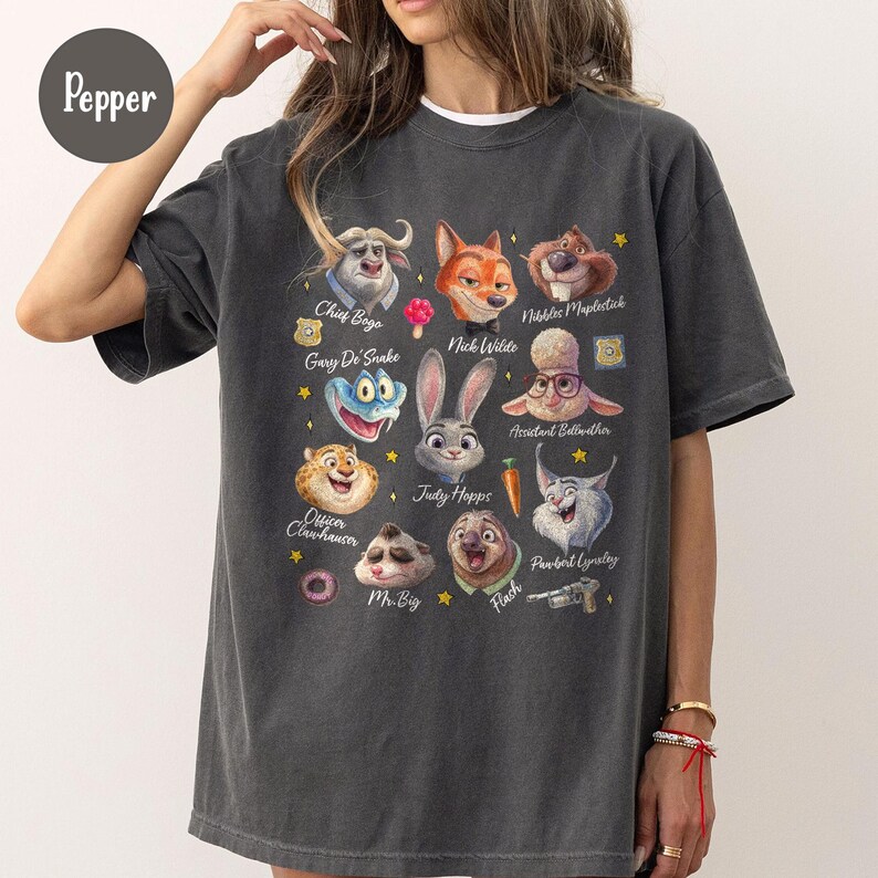 K&ouml;nnte beinhalten: Dunkelgraues T-Shirt mit farbenfrohen Cartoon-Charakterportr&auml;ts aus einem Film. Das Shirt zeigt Charaktere wie Judy Hopps, Nick Wilde und Chief Bogo. Der Text "Pepper" befindet sich in der oberen linken Ecke.