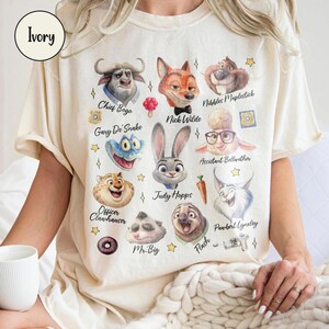 K&ouml;nnte beinhalten: Elfenbeinfarbenes T-Shirt mit Aquarellillustrationen von Zootopia-Charakteren, darunter Chief Bogo, Nick Wilde und Judy Hopps. Das Shirt hat einen Rundhalsausschnitt und kurze &Auml;rmel. Das Wort "Ivory" ist oben links aufgedruckt.