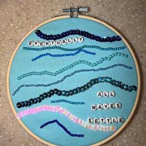 May include: A circular wooden embroidery hoop containing light blue fabric decorated with wavy lines of blue, teal, and iridescent pink sequins and beads. White plastic letter beads spell out the phrase EVENTUALLY ALL WAVES SETTLE.
