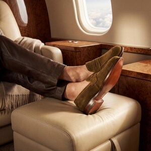 Mens feet resting on a leather ottoman in a private jet wearing premium olive brown suede loafers.