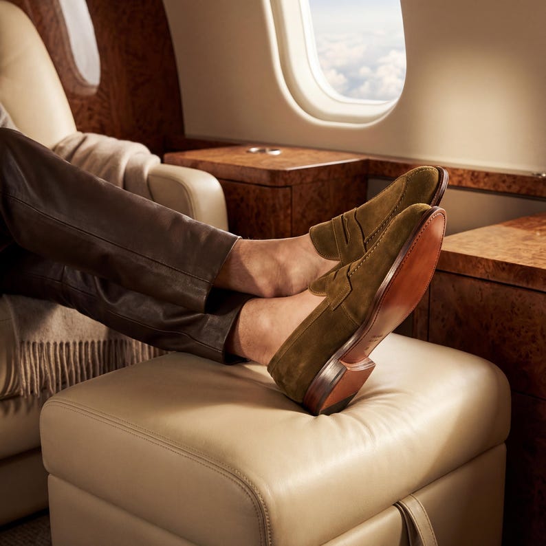 Mens feet resting on a leather ottoman in a private jet wearing premium olive brown suede loafers.