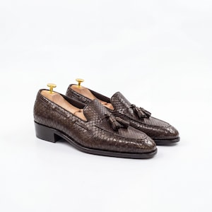 Könnte beinhalten: Ein Paar braune Leder-Loafer mit Schlangenmuster. Die Schuhe haben Quasten und einen kleinen Absatz. Die Loafer werden auf weißem Hintergrund präsentiert.
