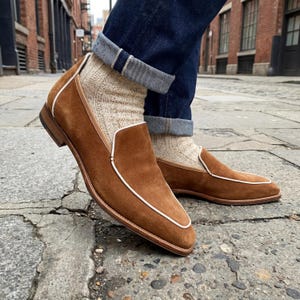 May include: Brown suede loafers with white trim. The shoes have a classic slip-on design and are paired with patterned socks and rolled-up blue jeans. The loafers appear to be made of high-quality materials.