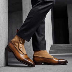 Hand-burnished leather dress boots with tan suede uppers leaning against a modern architectural concrete wall.