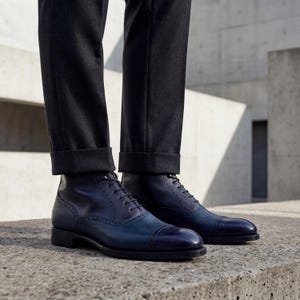 Two-tone navy blue leather boots styled with black formal trousers on a concrete background.