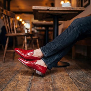 Close-up of red leather dress shoes being worn with dark denim jeans in a rustic cafe setting.
