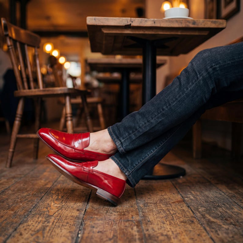 Close-up of red leather dress shoes being worn with dark denim jeans in a rustic cafe setting.