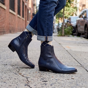 May include: Navy blue ankle boots with a textured, snakeskin-like pattern. The boots have a side zip and a low heel. The person is wearing blue jeans with the cuffs turned up, and argyle socks. The boots are on a city pavement.
