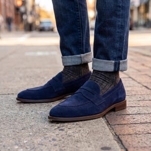May include: Navy blue suede penny loafers with brown soles. The shoes are paired with patterned gray socks and dark wash denim jeans with rolled cuffs. The image is taken outdoors on a brick and concrete surface.