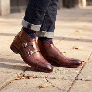 May include: Brown leather ankle boots with double buckle fastenings and red soles. The boots are worn with dark wash denim jeans and argyle socks. The boots appear to be made of high-quality leather.