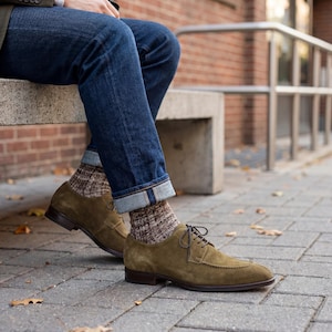 May include: Olive green suede dress shoes with dark brown soles and laces. The shoes are paired with patterned socks and blue denim jeans with rolled-up cuffs. The image is taken outdoors on a paved surface.