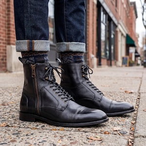 May include: Black leather boots with a cap toe, lace-up fastening, and side zip. The boots are worn with dark wash denim jeans and patterned socks. The boots appear to be new and unworn.
