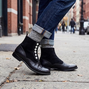 Pode incluir: Botas de tornozelo em couro preto e camurça com fecho de botão. As botas têm bico redondo e são combinadas com meias cinzentas estampadas e jeans azuis. As botas são de estilo clássico, adequadas para várias ocasiões.