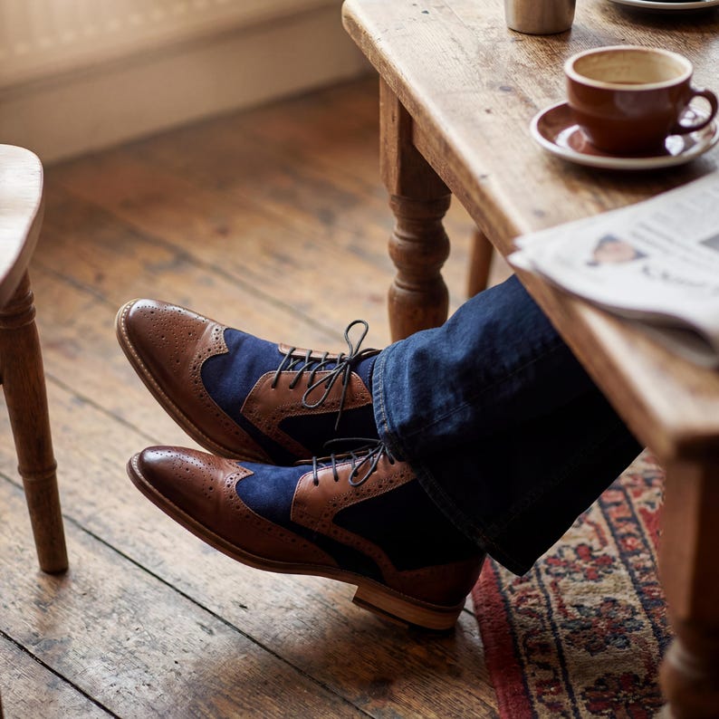 Overhead view of handcrafted leather boots with navy suede panels resting on a rustic wooden floor in a cafe.