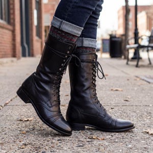 Puede incluir: Botas negras de cuero con cordones. Las botas tienen una caña alta, una puntera redondeada y un tacón bajo. Las botas se llevan con vaqueros oscuros y calcetines estampados. Las botas están sobre una superficie de hormigón.