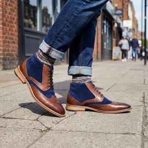 Low angle shot of a man wearing brown and navy wingtip boots with cuffed blue jeans on a city sidewalk.