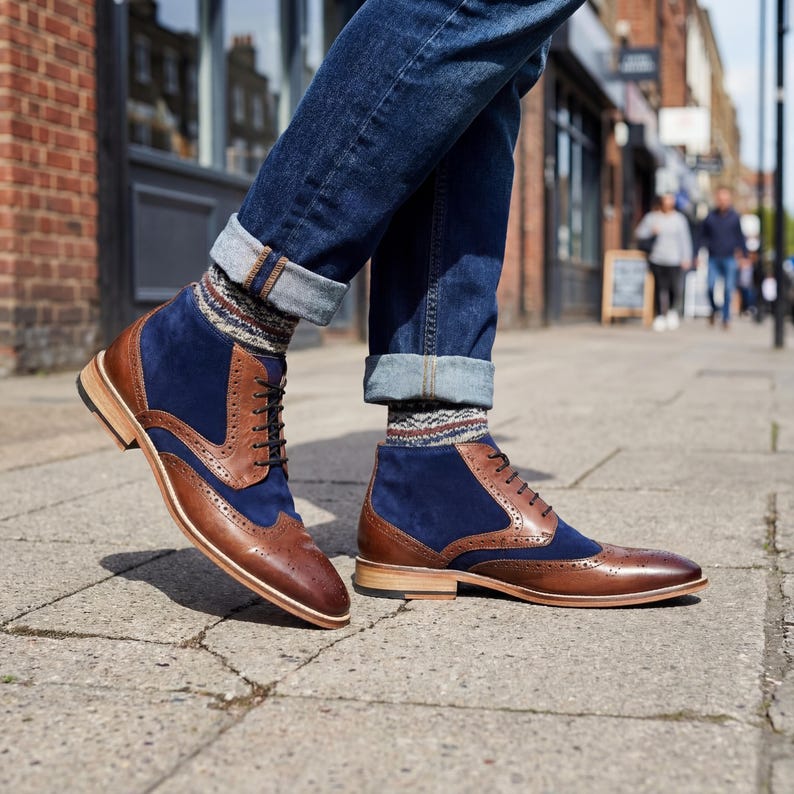 Low angle shot of a man wearing brown and navy wingtip boots with cuffed blue jeans on a city sidewalk.