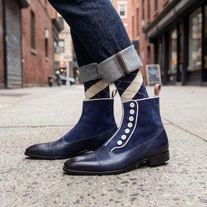 Pode incluir: Botas de camurça e couro azul marinho com detalhes de botões brancos e acabamento branco. As botas são combinadas com meias argyle e jeans escuros. Design clássico e elegante.
