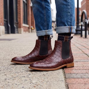 May include: A pair of brown leather Chelsea boots with a crocodile skin texture. The boots have a pointed toe, elastic side panels, and a low heel. The boots are worn with blue jeans and patterned socks.