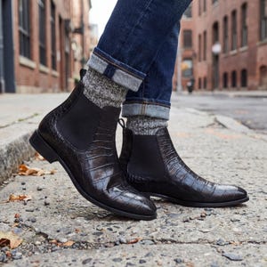 Puede incluir: Botas Chelsea negras con un patrón de textura de cocodrilo. Las botas tienen paneles laterales elásticos y un tacón bajo. La persona lleva vaqueros azules y calcetines grises. Las botas están en una acera.