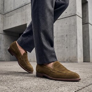 Man walking in grey flannel trousers and handmade brown suede slip-on shoes on concrete.