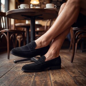 May include: A pair of black suede loafers with a classic penny strap design. The shoes are displayed indoors, with a wooden floor and a blurred background of a cafe setting. The loafers have a low heel and a sleek, minimalist aesthetic.
