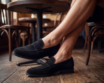 May include: A pair of black suede loafers with a classic penny strap design. The shoes are displayed indoors, with a wooden floor and a blurred background of a cafe setting. The loafers have a low heel and a sleek, minimalist aesthetic.