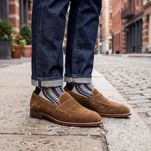 Man wearing luxury tobacco suede loafers with dark denim jeans on a cobblestone city street.