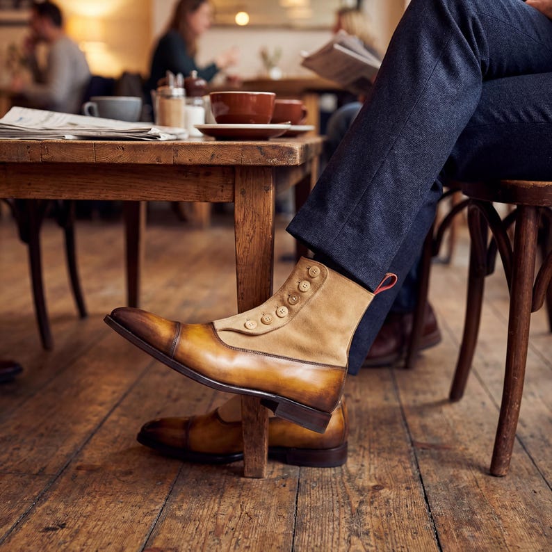 Luxury mens spat boots in amber brown paired with navy wool trousers in a sophisticated cafe setting.