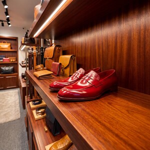 Pair of high-shine oxblood red mens loafers displayed on a dark wood boutique shelf with leather accessories.