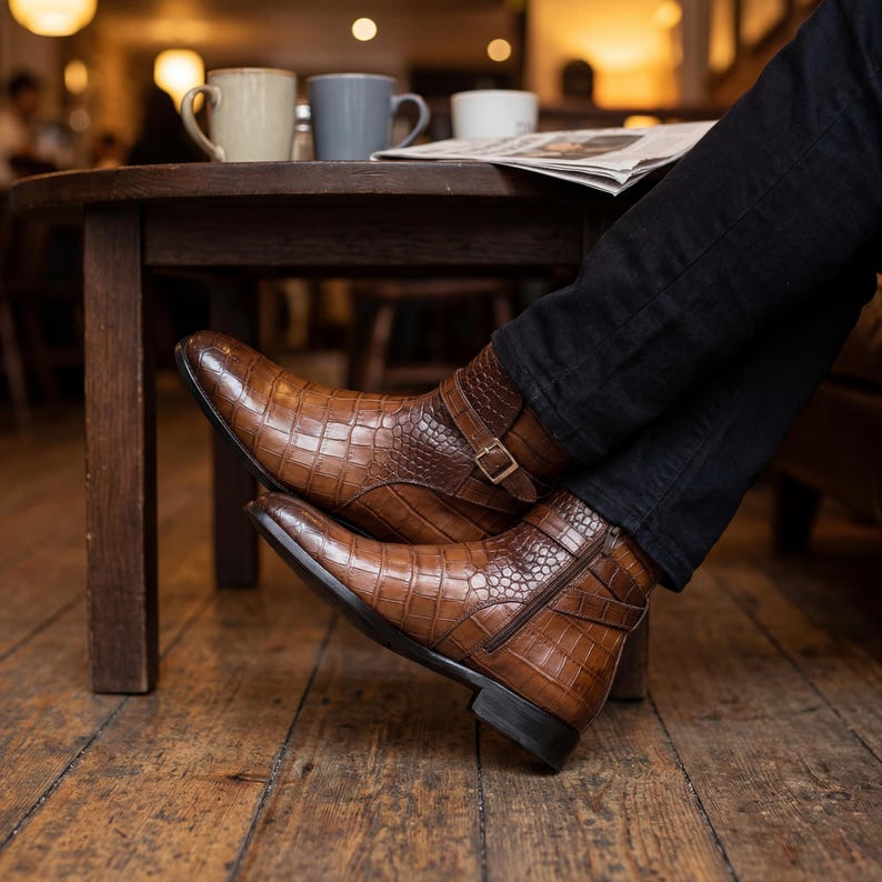 Men's exotic leather zip-up boots styled with black denim in a cozy upscale cafe setting.
