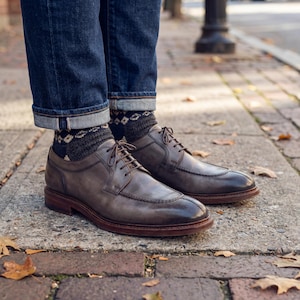May include: A pair of gray leather dress shoes with brown soles and laces. The shoes are worn with patterned socks and dark blue jeans. The image is taken outdoors on a brick walkway.