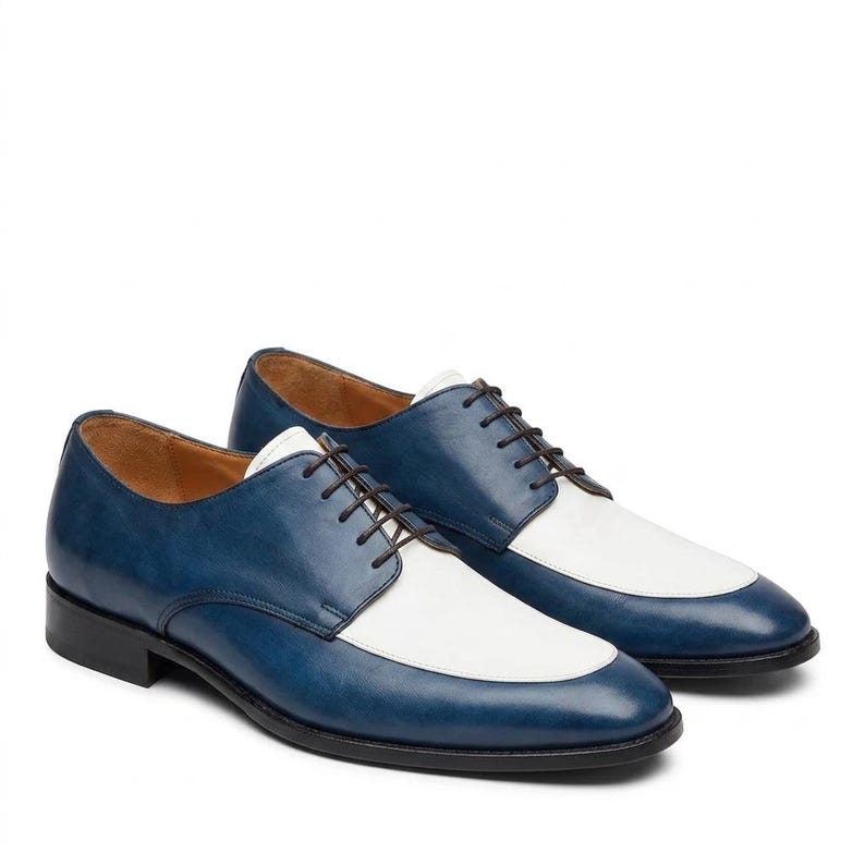 May include: A pair of two-tone oxford shoes featuring a deep blue leather finish on the sides and heel, contrasted by a smooth white leather vamp. These lace-up shoes include thin dark brown laces, a low black heel, and a tan interior lining.