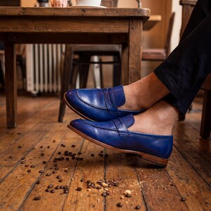 Men's blue leather slip-on shoes worn without socks with navy trousers in a rustic cafe setting.