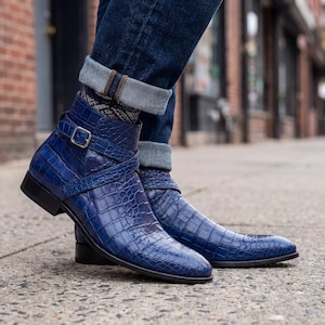 May include: A pair of blue leather ankle boots with a crocodile skin texture. The boots have a strap with a silver buckle and a black sole. The boots are worn with blue jeans.