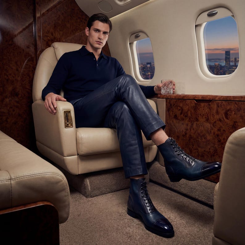 Peut inclure: Un homme portant un polo bleu marine, un pantalon en cuir et des bottines en cuir bleu &agrave; lacets, assis dans un fauteuil en cuir cr&egrave;me &agrave; l'int&eacute;rieur d'un jet priv&eacute;. Les bottes ont un bout fonc&eacute;. Un verre de liquide est dans sa main.