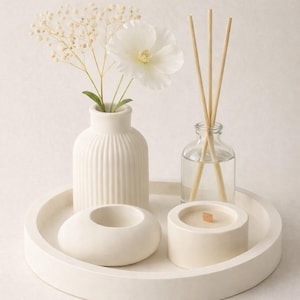 Puede incluir: Una bandeja decorativa blanca contiene un pequeño jarrón blanco con una flor blanca y ramitas, una botella de vidrio transparente con varillas de madera, un pequeño portavelas blanco y una vela blanca con una mecha de madera. Los artículos están dispuestos sobre una superficie blanca.