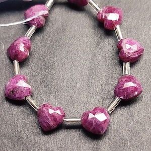 Könnte beinhalten: Eine Kette aus herzförmigen, facettierten Rubinperlen. Die tiefmagentafarbenen Edelsteine sind auf einen silberfarbenen Draht gefädelt. Am Ende der Kette ist eine weiße Quaste befestigt. Die Perlen haben einen Durchmesser von ca. 1,3 cm.
